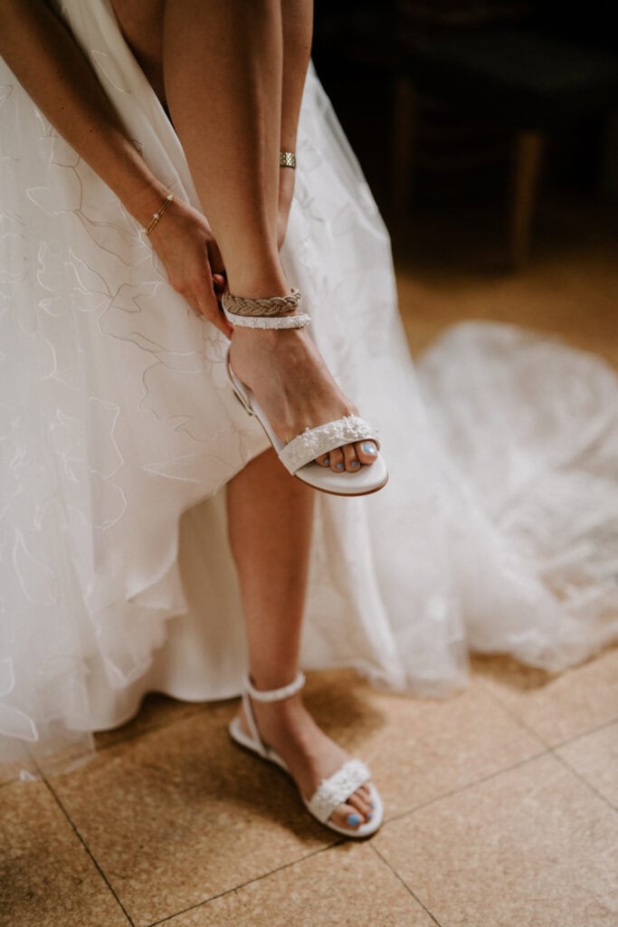 At a charming Kent wedding venue, a bride adjusts the ankle strap of her white sandal while lifting her wedding dress. The sandals have decorative elements, and her toenails are painted light blue. She wears a delicate bracelet on her wrist, and the gown features a textured pattern. Image by Pearce Wedding Photography.
