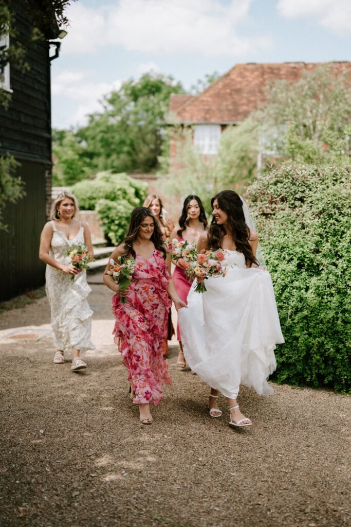 A bride in a white wedding dress walks outside with four bridesmaids, each in different pink dresses, holding bouquets of flowers. They are on a gravel path at a Kent wedding venue, surrounded by greenery and buildings, on a sunny day. Everyone is smiling and enjoying the moment. Image by Pearce Wedding Photography.