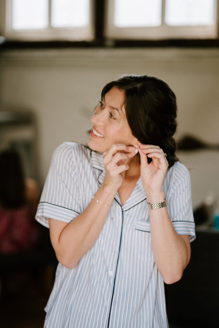 A woman with dark hair smiles while putting on an earring. She is wearing light blue and white striped pajamas and a watch. The background, reminiscent of beautiful Kent wedding venues, is softly blurred, highlighting her centered in the frame. Natural light from a window brightens the scene. Image by Pearce Wedding Photography.