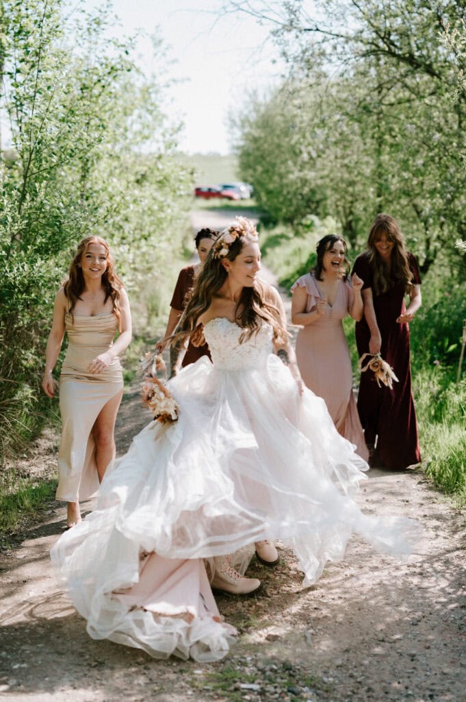 A bride in a flowing white dress, adorned with a floral crown, walks joyfully along a sunlit path at a picturesque Kent wedding venue with her bridesmaids. The bridesmaids wear dresses in various shades of pink and beige, and hold bouquets. Trees and greenery border the path, creating a serene, natural setting. Image by Pearce Wedding Photography.