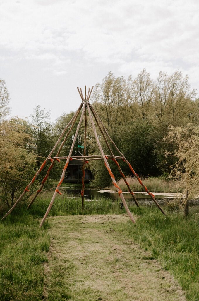 A tipi frame made of wooden poles tied together with orange cloths stands on a grass path in the wilderness. Surrounding the structure are trees with light green foliage and a cloudy sky overhead, creating an enchanting scene perfect for a Canterbury wedding photographer to capture. Image by Pearce Wedding Photography.