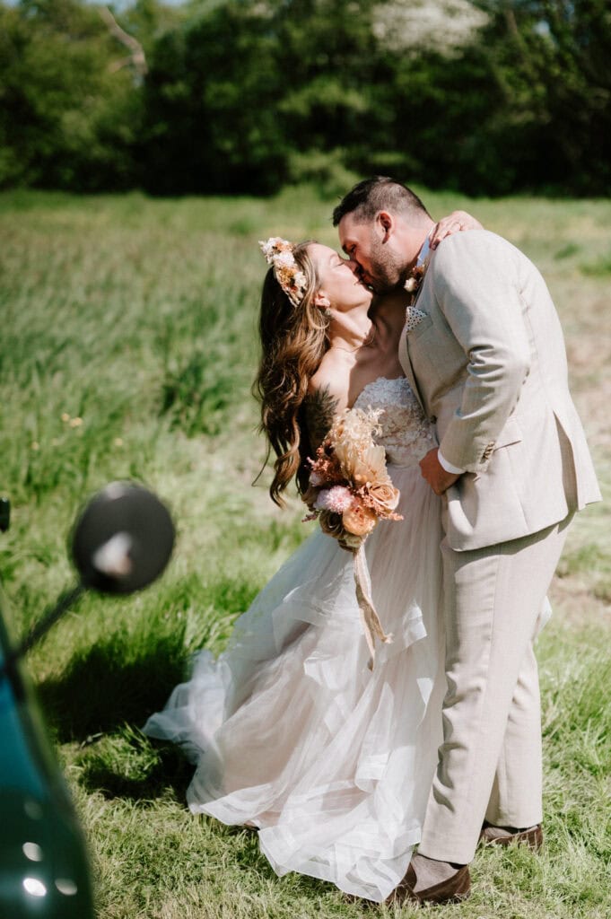A bride and groom share a passionate kiss outdoors in a grassy field. The bride wears a strapless white gown and holds a bouquet, while the groom is dressed in a light-colored suit. Captured by their Canterbury wedding photographer, the sun shines brightly, illuminating the greenery around them. Image by Pearce Wedding Photography.