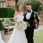 A bride and groom standing outdoors at a charming Kent wedding venue, with the bride in a white off-the-shoulder lace wedding dress holding a bouquet of flowers, smiling, and the groom in a green plaid suit showing off the lining of his jacket with a playful expression. They are in a garden setting. Image by Pearce Wedding Photography.