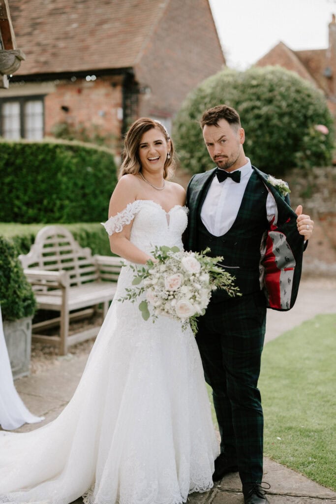 A bride in a lace off-shoulder gown holding a bouquet stands next to a groom in a checkered suit showing the red lining of his jacket. They are outdoors in a garden with greenery and a brick building, capturing the charm of a Kent wedding amidst the timeless beauty of winters barns.