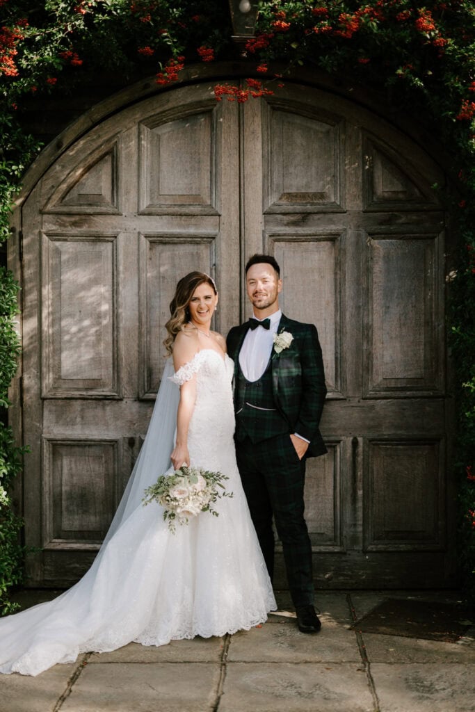 A bride and groom stand together in front of a large wooden door at Winters Barns. The bride holds a bouquet and wears an off-the-shoulder wedding dress; the groom wears a dark suit with a vest and bow tie, ready to celebrate their Kent wedding.