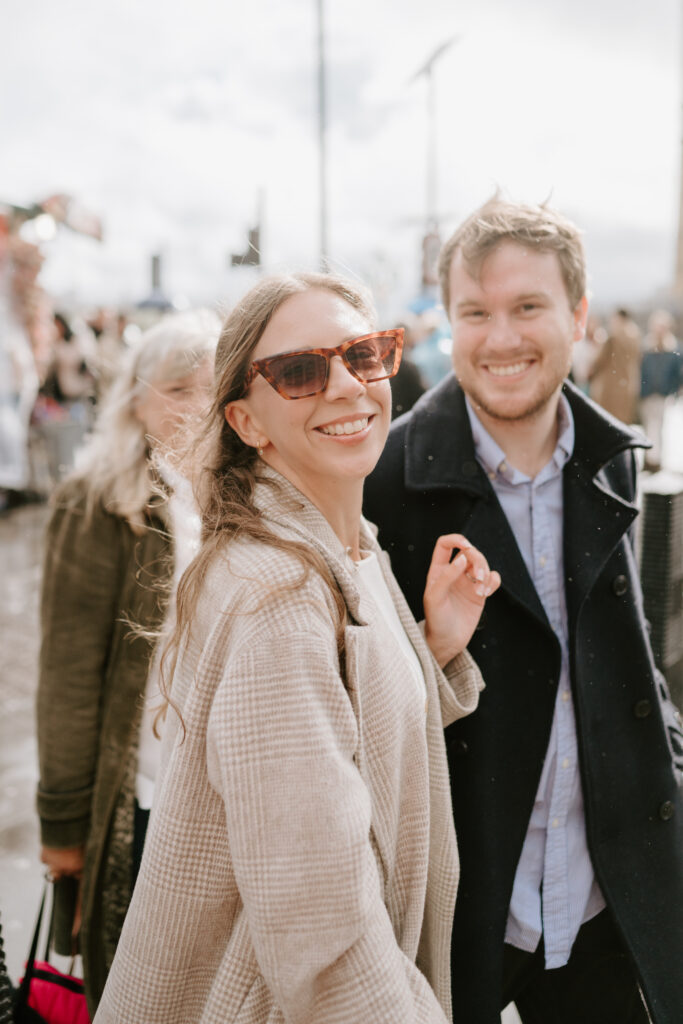 A woman wearing sunglasses and a checkered coat smiles while standing next to a man in a dark coat. Captured by a candid photographer, they are outdoors on a bustling street, with other people in the background. The sun is shining brightly. Image by Pearce Wedding Photography.