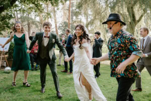 A joyful group, captured by a candid wedding photographer, including a bride in a white dress and a groom in a green suit, are holding hands and dancing on the grassy lawn. One man sports a floral shirt and hat. Trees and guests frame the cheerful scene in the background. Image by Pearce Wedding Photography.