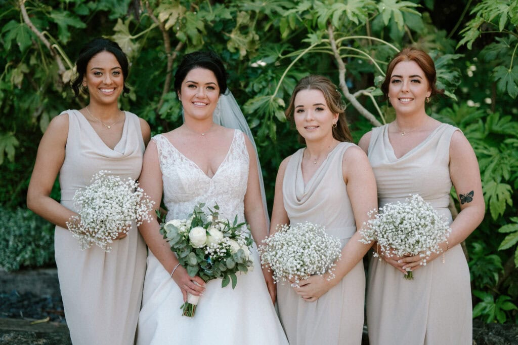 Four women in light taupe dresses posing together outdoors, with three holding bouquets of baby's breath and the fourth, a bride in a white dress with a veil, holding a bouquet of white flowers and greenery. Lush green foliage is in the background. They are smiling, captured perfectly by our Maidstone wedding photographer. Image by Pearce Wedding Photography.
