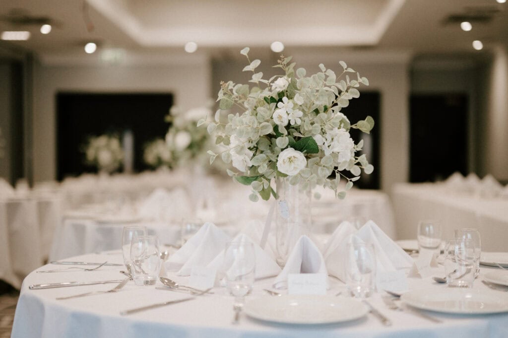 A beautifully set dining table at the Village Hotel, adorned with a centerpiece of white flowers and greenery. The table is covered with a white tablecloth and arranged with white napkins, glassware, and cutlery, creating an elegant and sophisticated ambiance in a well-lit room—perfect for any Maidstone wedding photographer to capture. Image by Pearce Wedding Photography.