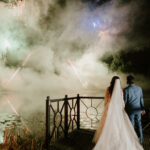 A couple in wedding attire stands on a wooden platform by a lake at night, captured perfectly by their Kent wedding photographer as fireworks light up the sky. Smoke and reflections create a dreamy atmosphere around them. Image by Pearce Wedding Photography.