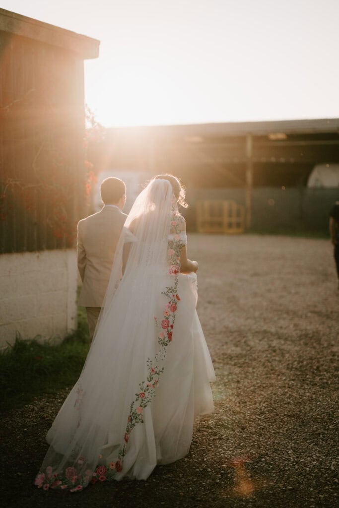 A bride and groom walk into the sunset on a gravel path, captured by a candid wedding photographer. The bride wears a flowing white dress with floral embroidery and a veil, while the groom dons a light suit. They are surrounded by rustic buildings and soft sunlight. Image by Pearce Wedding Photography.