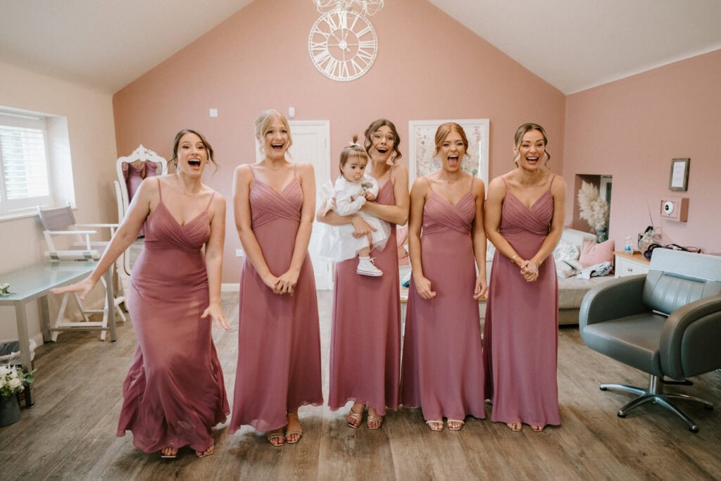 A group of six women, five in matching mauve dresses, stand in a room with pale pink walls. One woman holds a small child in a white outfit. They are all smiling with expressions of joy and excitement, captured perfectly by the Canterbury wedding photographer at Winters Barns. The room features minimalistic decor and a modern clock on the wall. Image by Pearce Wedding Photography.
