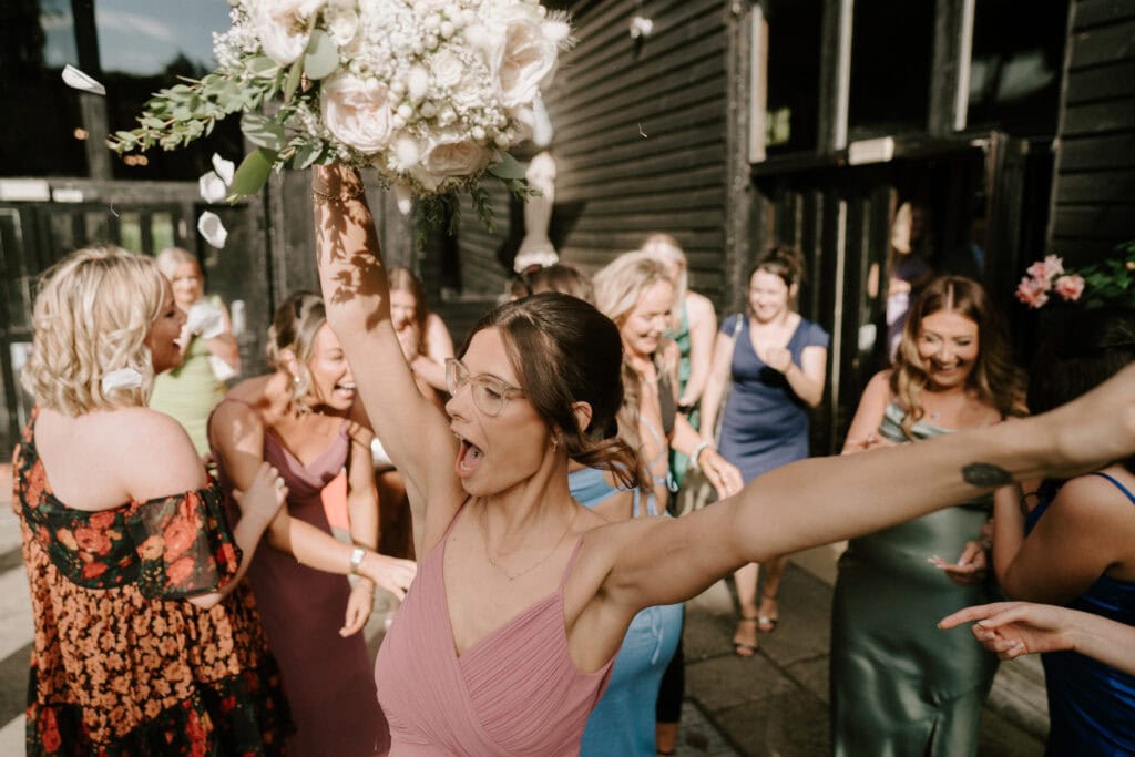 A joyful woman in a pink dress raises her arms while holding a bouquet of flowers, celebrating with a group of women outside. Everyone is smiling and dressed in colorful outfits, indicating a festive mood. Sunlight illuminates the scene, enhancing the lively atmosphere—captured brilliantly by the Canterbury wedding photographer. Image by Pearce Wedding Photography.