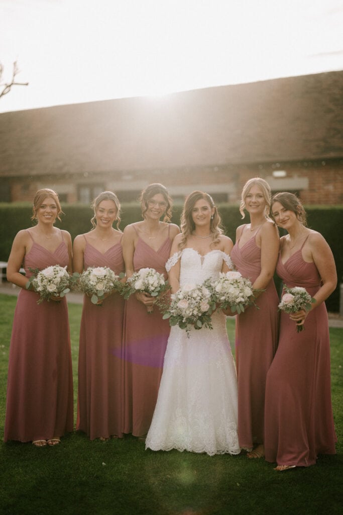 A bride stands outdoors with five bridesmaids at Winters Barns. The bride wears a white lace wedding dress and holds a bouquet of flowers. The bridesmaids, dressed in matching mauve gowns, also hold bouquets. They are all smiling with a bright sunlit sky and a charming building in the background. Image by Pearce Wedding Photography.