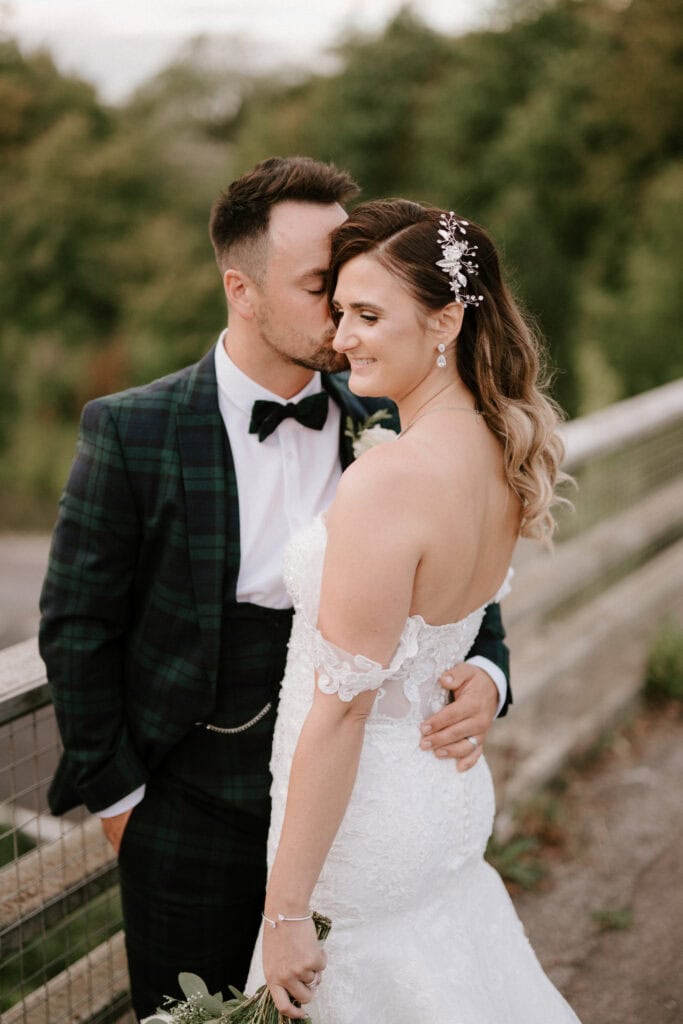 A couple stands outdoors on their wedding day. The groom, in a dark tartan suit and bow tie, leans in to kiss the bride's forehead. The bride, in a lace off-shoulder white gown with a floral hair accessory, smiles softly. They are standing in front of a wooden fence at Winters Barns, beautifully captured by a Canterbury wedding photographer. Image by Pearce Wedding Photography.