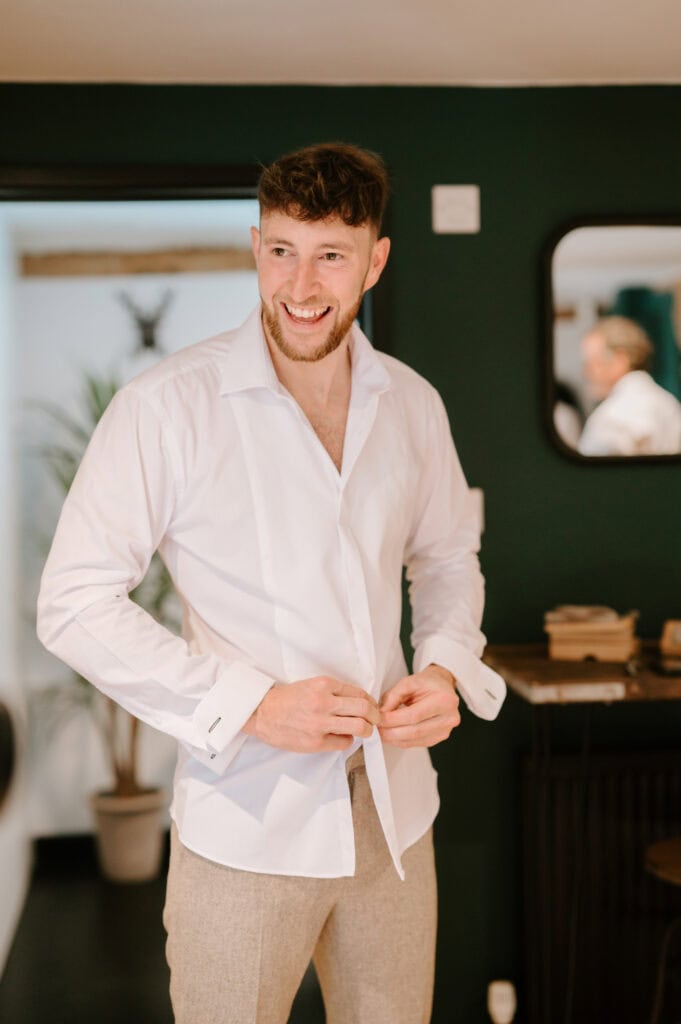 Man buttoning white shirt and smiling indoors.