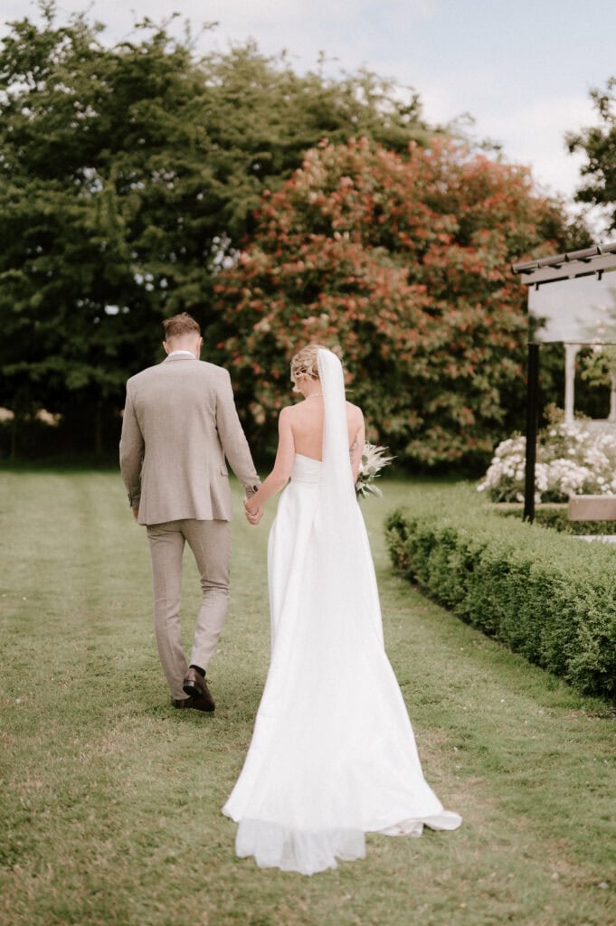 A bride and groom walk hand in hand down a grassy path, the bride's long white dress and veil trailing behind her. The groom is dressed in a beige suit. They are surrounded by lush greenery and flowers, with a cloudy blue sky in the background, captured perfectly by their Dover wedding photographer. Image by Pearce Wedding Photography.