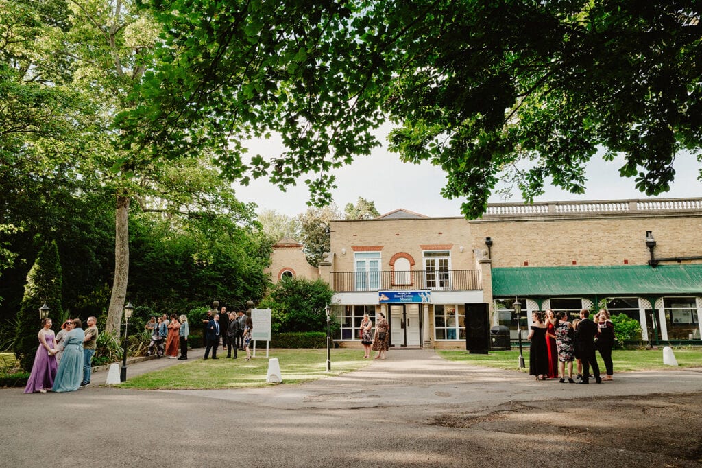 A group of elegantly dressed people gathers outside a picturesque Thanet wedding venue, surrounded by lush greenery. The large brick building features arched windows and a canopy, perfect for capturing stunning photographs on this sunny day. Image by Pearce Wedding Photography.