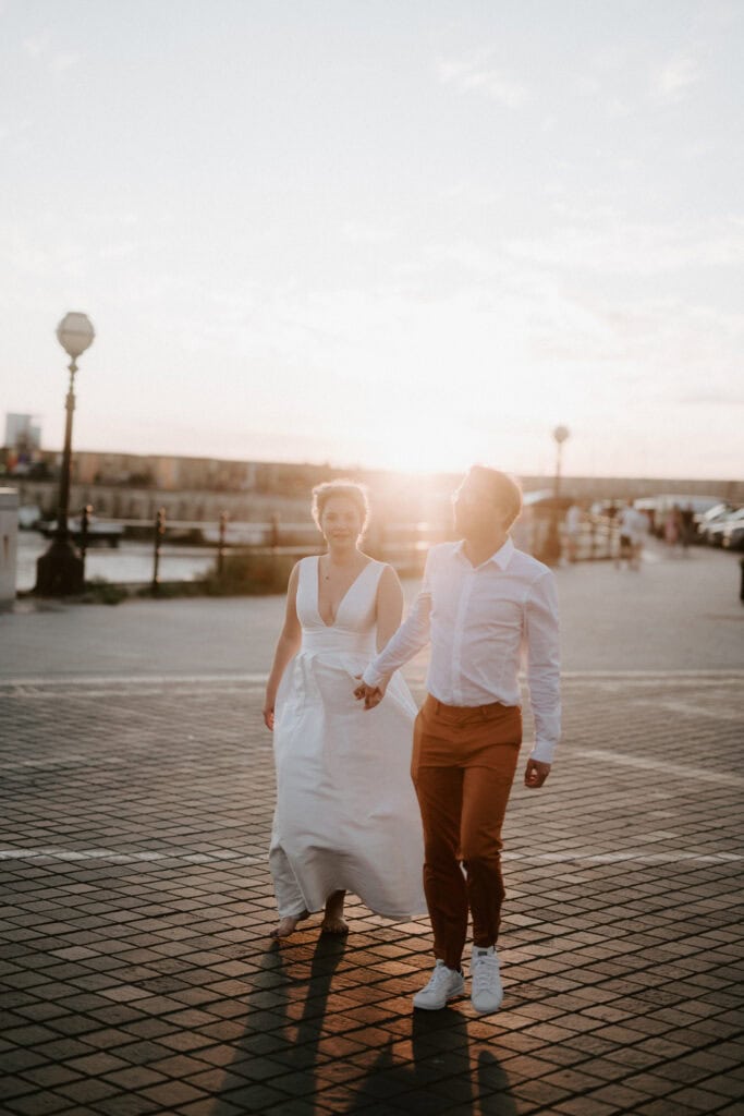 A couple walks hand in hand along a sunlit boardwalk, embodying the essence of a Thanet wedding. The woman wears a long white dress, and the man is in a white shirt and brown pants. The sun sets in the background, casting a warm glow over the enchanting scene. Image by Pearce Wedding Photography.