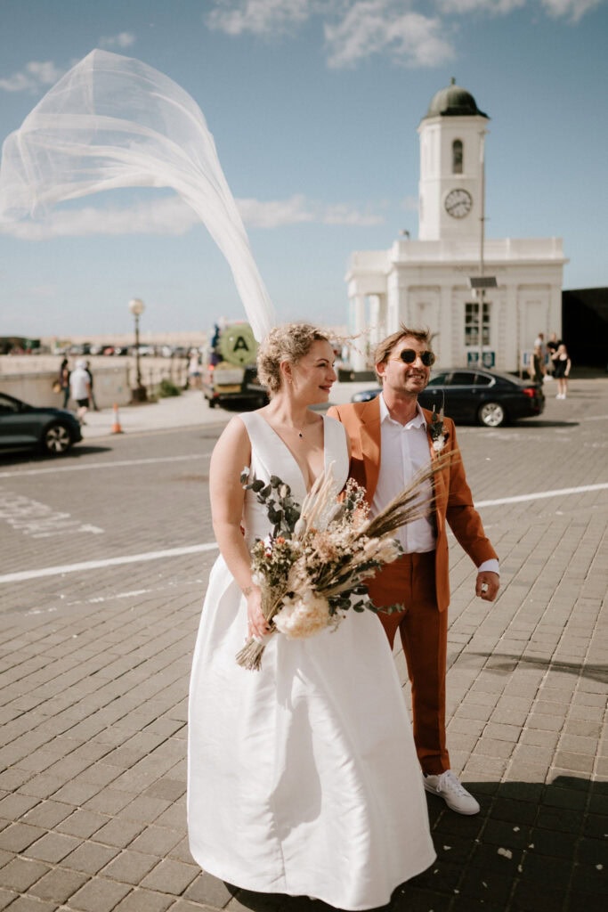 A couple stands smiling outdoors on a sunny day at a picturesque Thanet wedding venue. The woman wears a white dress and holds a bouquet, her veil flowing in the wind. The man sports an orange suit. They stand on a paved area near a building with a charming clock tower. Image by Pearce Wedding Photography.