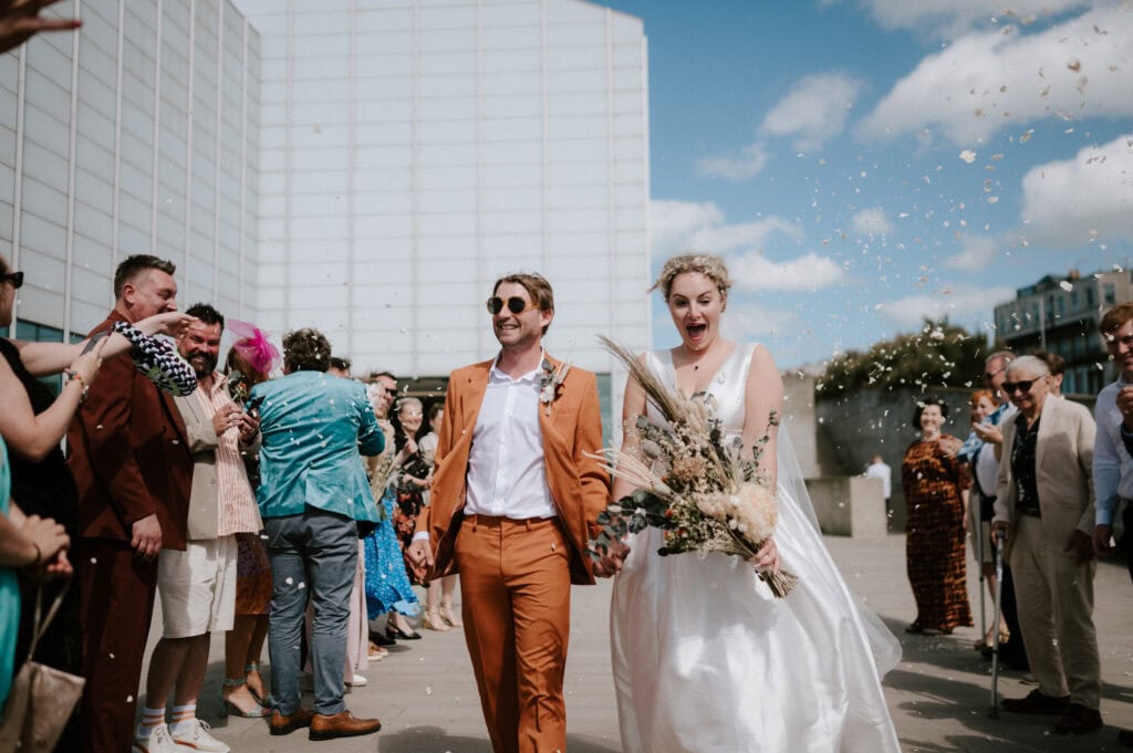 A joyful couple walks hand in hand down the aisle, exuding happiness in their wedding attire. The groom sports an orange suit, while the bride dazzles in white with a bouquet. Guests cheer and throw confetti against the backdrop of a modern building and blue sky—a perfect scene for any Thanet wedding venue. Image by Pearce Wedding Photography.