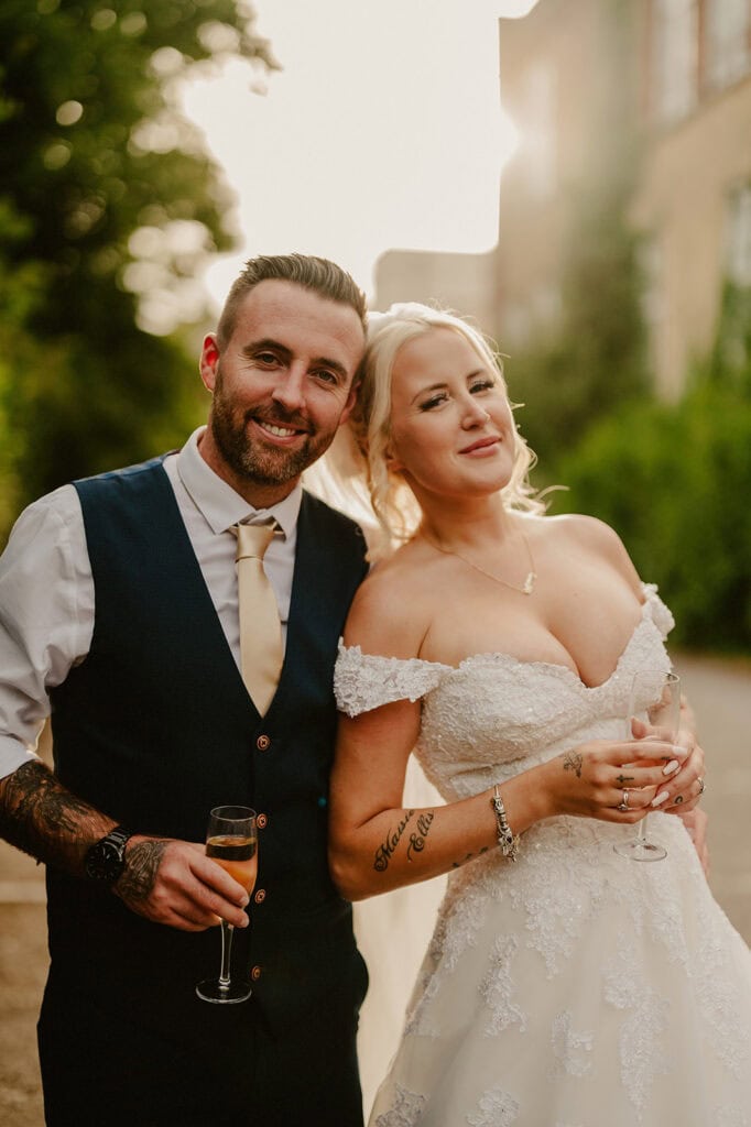 A couple in wedding attire smiles warmly, captured beautifully by a Thanet wedding photographer. The groom wears a dark vest and tie, while the bride stuns in an off-shoulder white gown. Both hold champagne glasses, posed against a softly blurred backdrop of greenery and a building, perhaps at a Thanet wedding venue. Image by Pearce Wedding Photography.