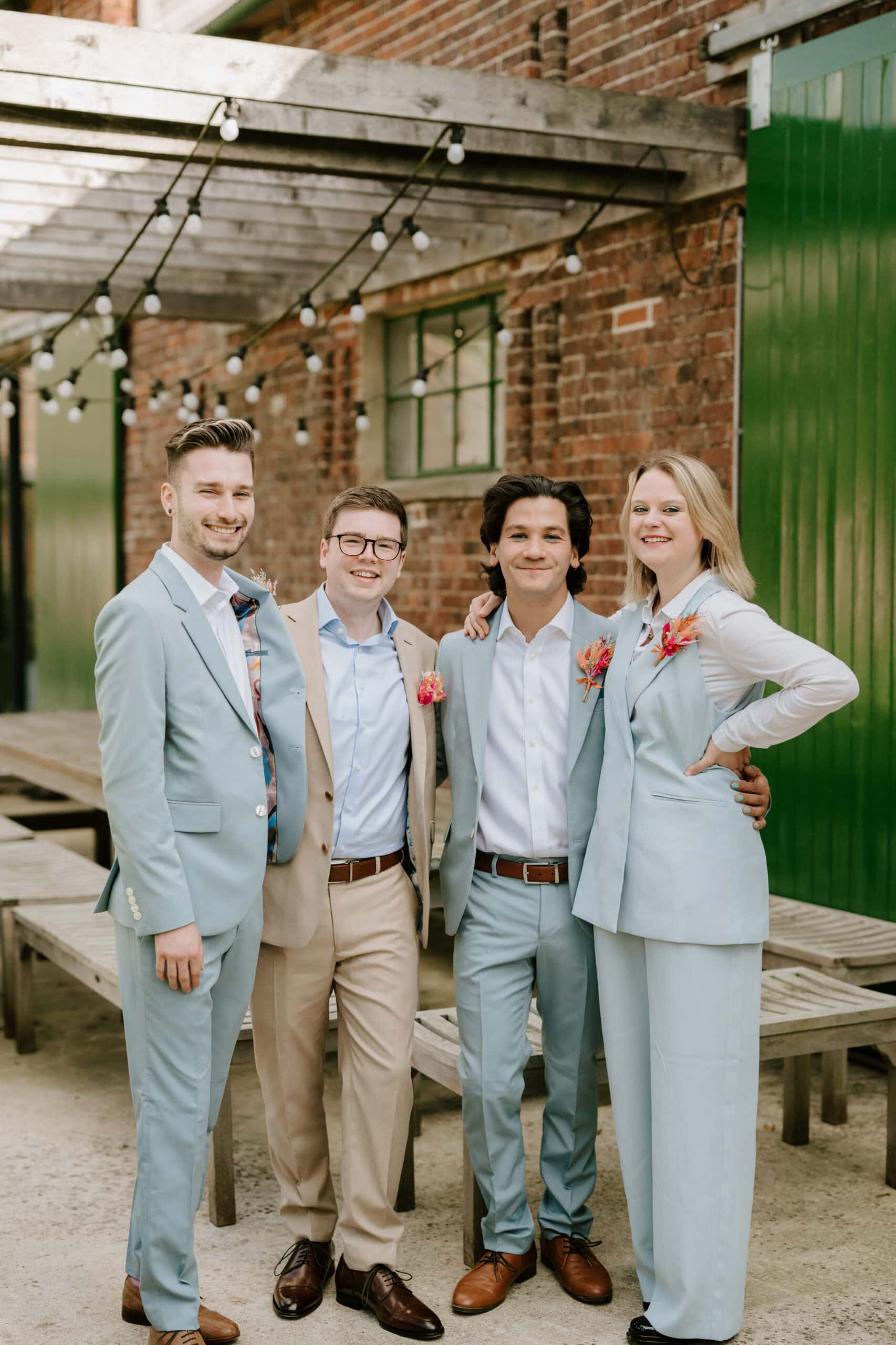 Four people smiling and posing outdoors against a brick wall and green doors. Three are wearing light blue suits, while one is in a tan suit. Captured by a Kent wedding photographer, they stand under a pergola with string lights, beside wooden benches. Image by Pearce Wedding Photography.