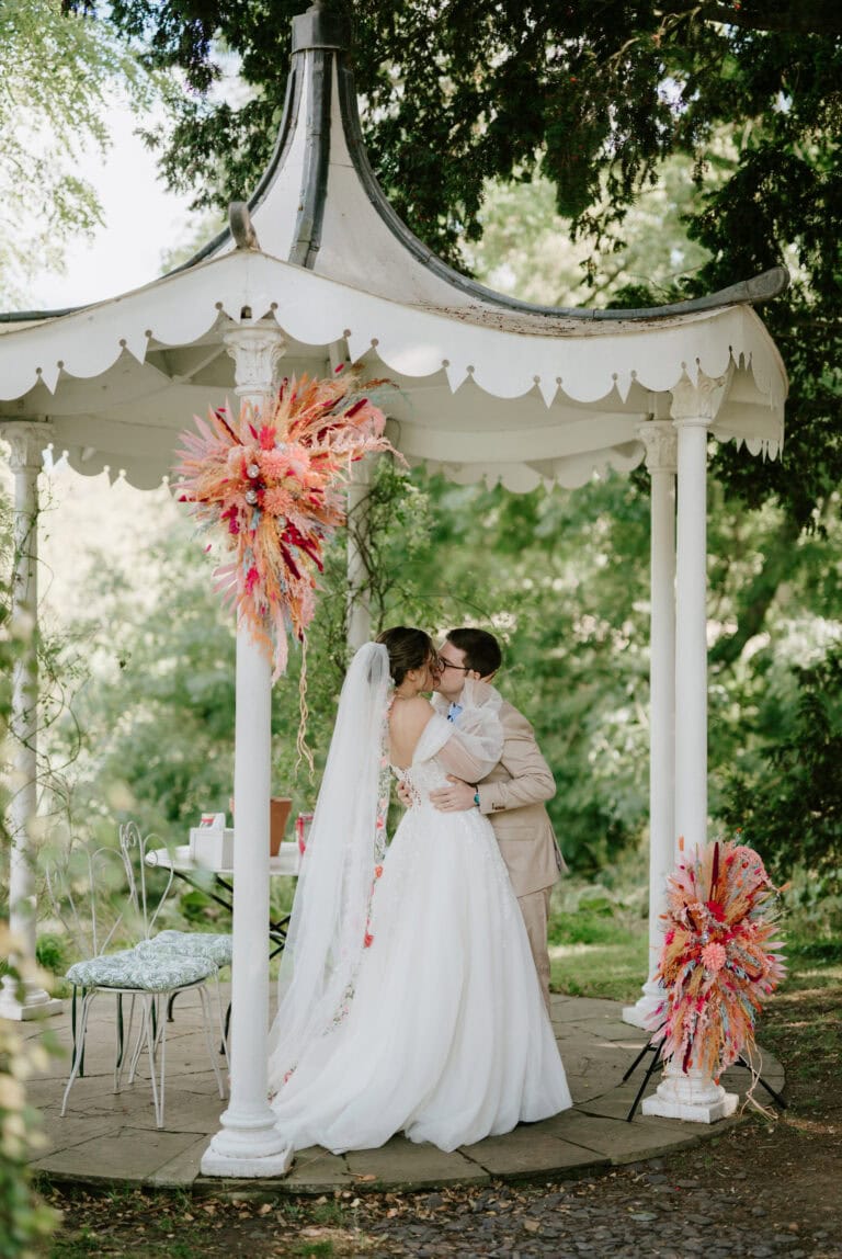 Under a white gazebo at Preston Court, adorned with vibrant floral arrangements, a couple shares a kiss. The bride’s flowing gown and veil complement the groom's light suit, while lush greenery completes this romantic Kent wedding venue setting. Image by Pearce Wedding Photography.