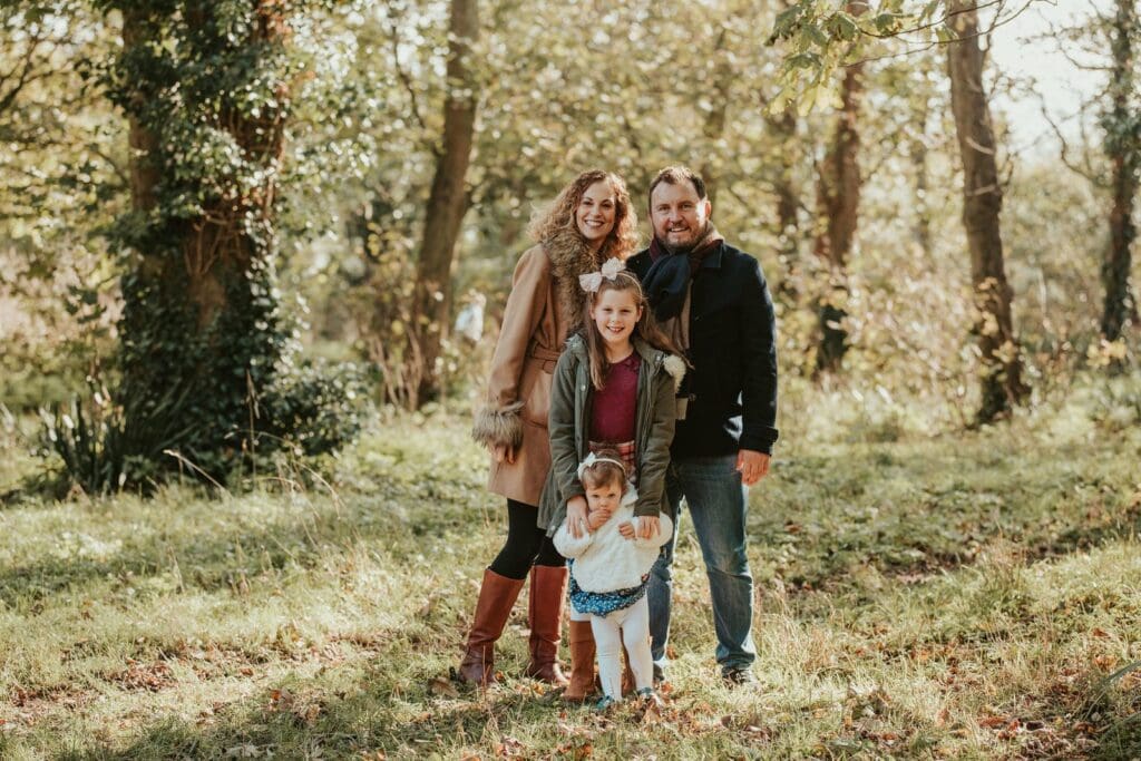 A smiling family of four poses outdoors in a wooded area, with the parents standing behind their two young daughters.