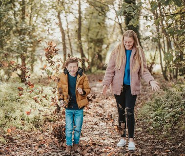 A family strolls through a leaf-covered path in a forest near Thanet, enjoying the autumn weather. The boy is tossing leaves in the air while the woman smiles, all captured beautifully by a photographer.