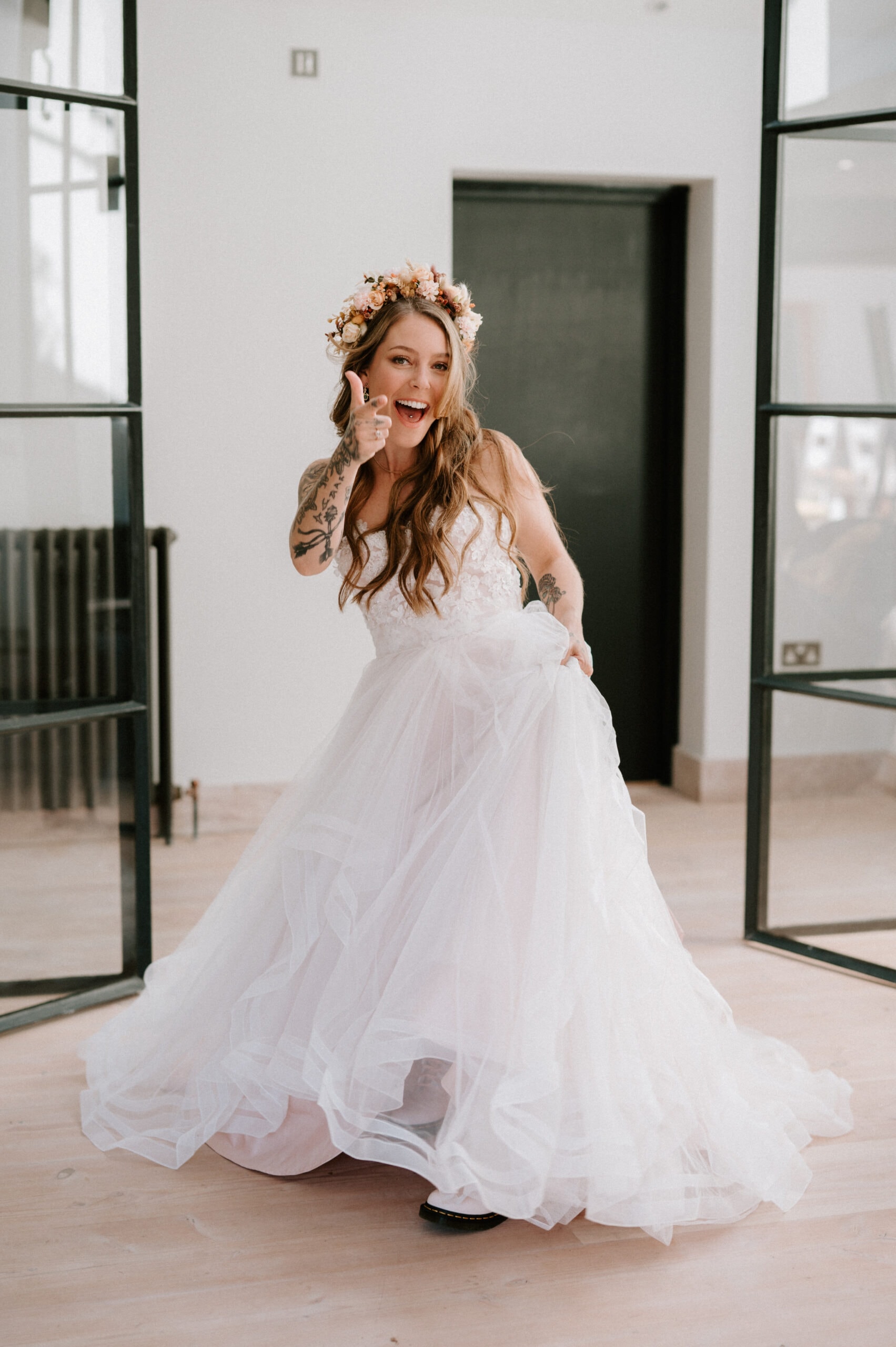 A bride dressed in a white wedding gown and floral headpiece poses playfully in a modern, well-lit room with black-framed glass doors, captured beautifully by her wedding photographer.