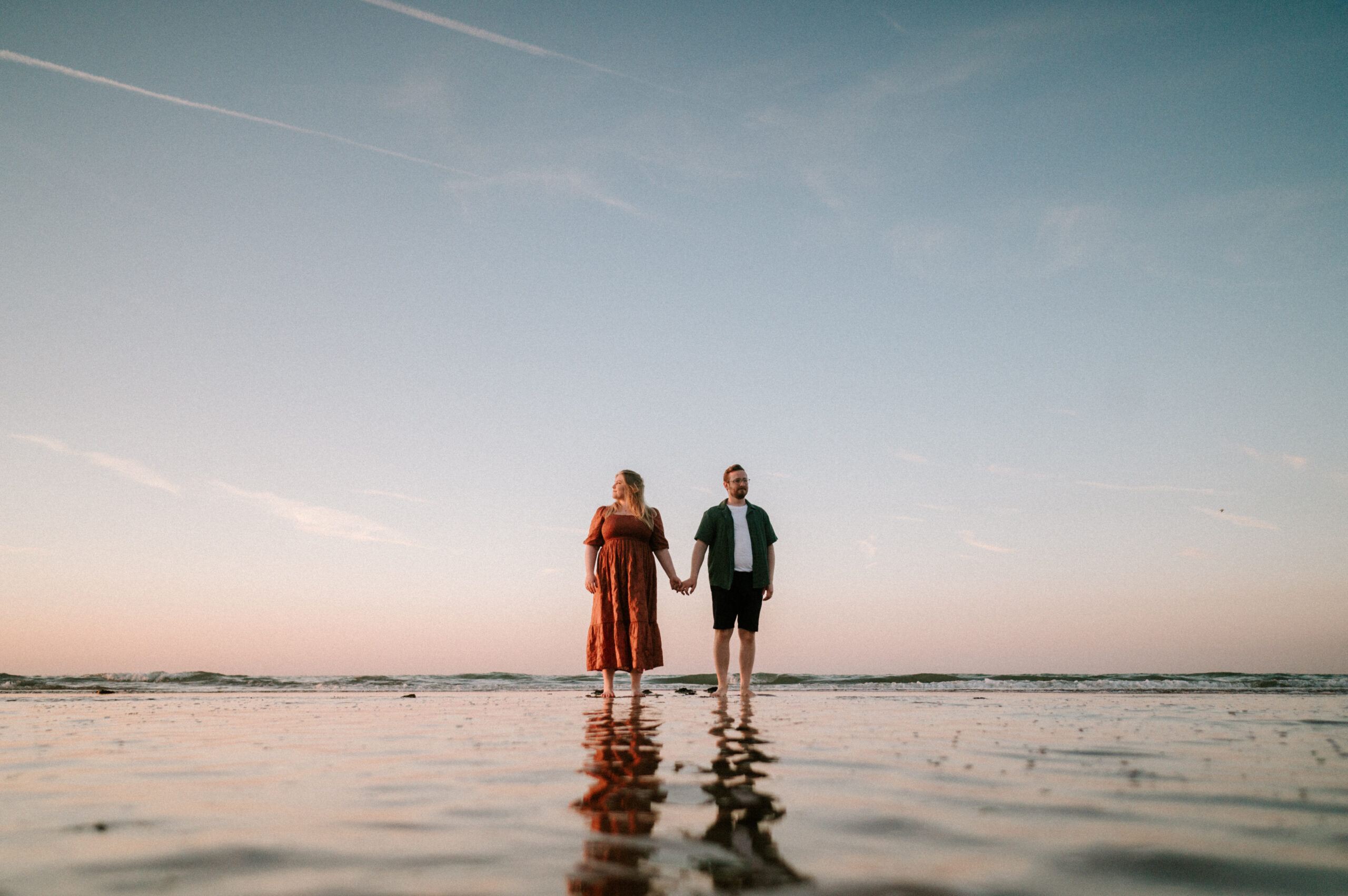 A couple holds hands while standing on a shallow beach at sunset in Kent, with clear skies and thin clouds above, perfectly captured by their wedding photographer.