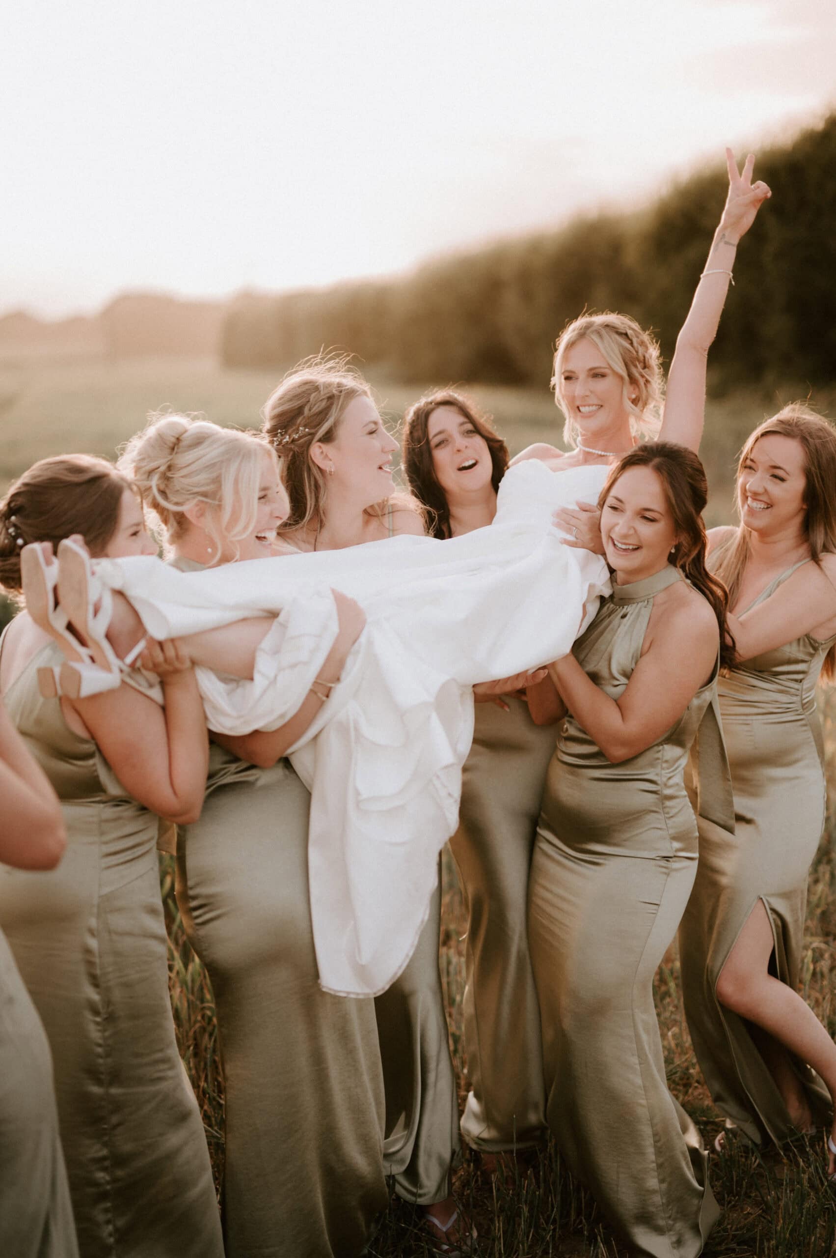 A group of women in matching light green dresses hold up a woman draped in a white dress, smiling and celebrating in an outdoor setting, captured beautifully by a wedding photographer in Kent.