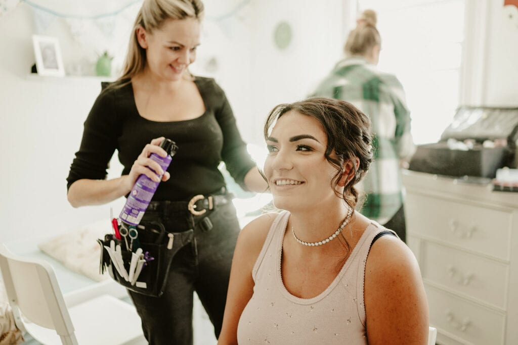 A woman in a pink tank top sits in a chair smiling as another woman in a black shirt styles her hair with hairspray. Another person is blurred in the background.