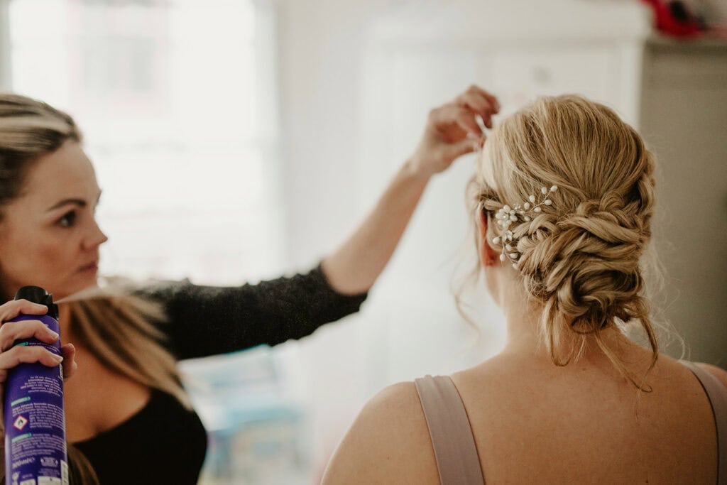A woman stylist sprays the hair of another woman whose back is turned, styling her intricate updo adorned with small flowers.
