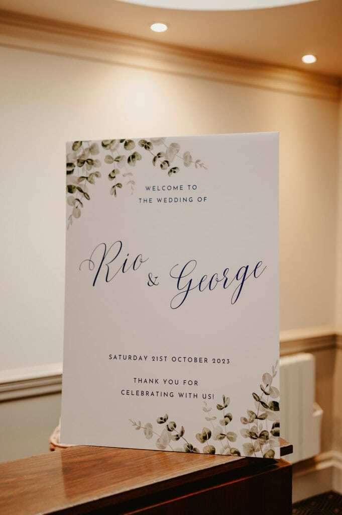Wedding sign that reads, "Welcome to the wedding of Rio & George, Saturday 21st October 2023. Thank you for celebrating with us!" The sign features a floral design.