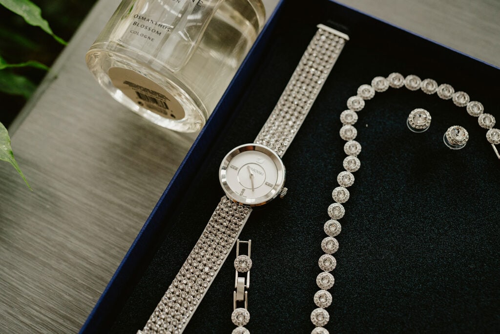 A silver watch, bracelet, necklace, and earrings with clear gemstones are placed in a black jewelry box next to a bottle of Osmanthus Blossom cologne.