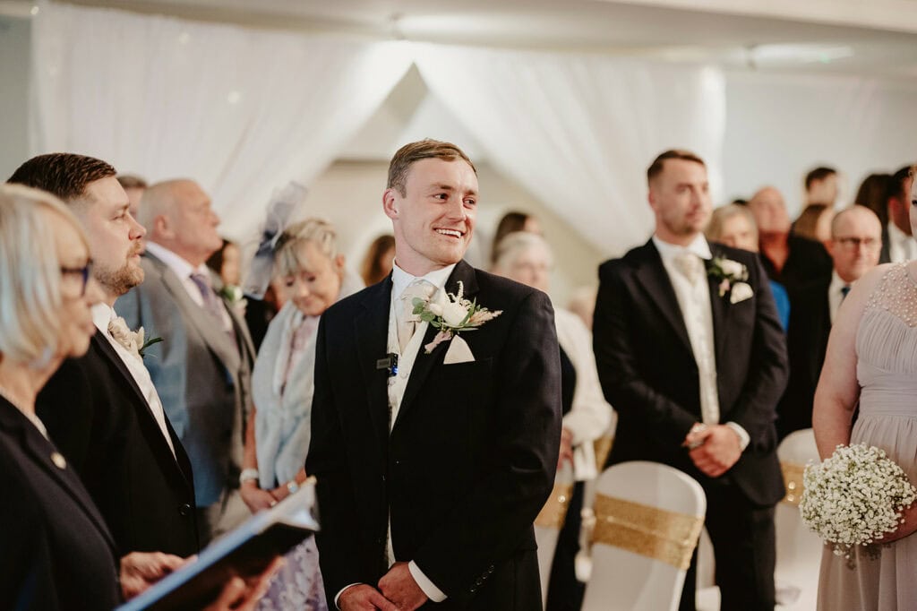 A man in a suit, smiling, stands among wedding guests indoors. Other attendees appear attentive, with some holding flowers. White drapery decorates the background.