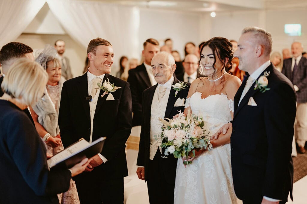 A bride and groom stand at their wedding ceremony with the bride's father and another older man, surrounded by guests.