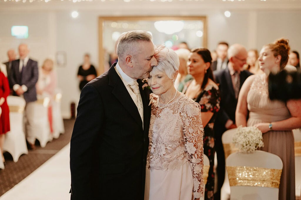 A groom in a black suit kisses an elderly woman on the forehead during a wedding ceremony. Guests are seated on white and gold chairs in the background.