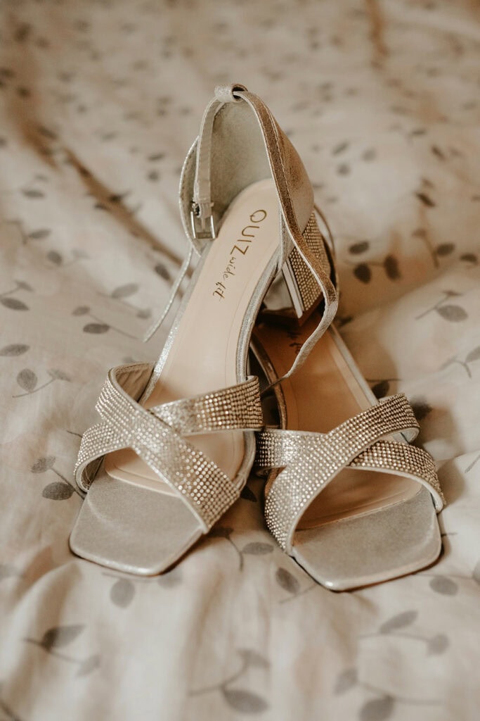 A pair of silver, open-toe high-heeled shoes with crisscross straps adorned with rhinestones, placed on a patterned fabric surface.