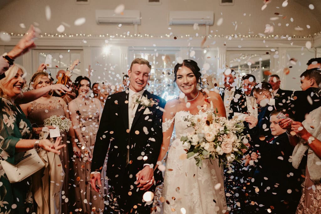 A bride and groom smile and hold hands while walking through a crowd of guests throwing confetti indoors. The bride holds a bouquet, and both are dressed in wedding attire.
