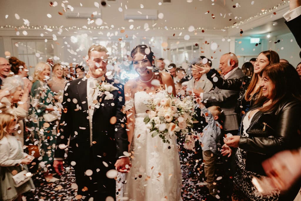A newly married couple holds hands and smiles while walking through a crowd of guests throwing confetti inside a decorated room at the elegant Abode Hotel Canterbury. The bride carries a bouquet of flowers, capturing the perfect wedding moment.