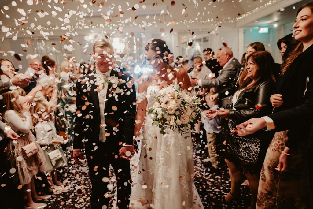A newly married couple walks down the aisle amid a shower of confetti, holding hands and smiling, surrounded by cheering wedding guests.