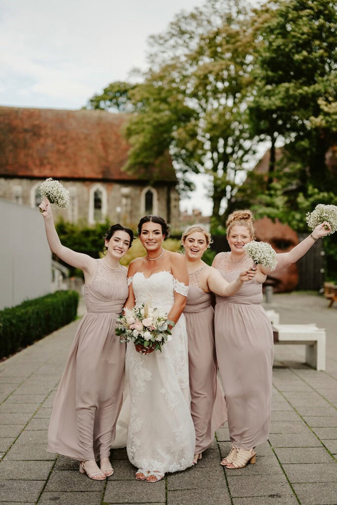 A bride in a white dress stands with three bridesmaids in matching beige dresses, all holding bouquets. They are outdoors, in front of a church. The bridesmaids raise their arms in celebration.