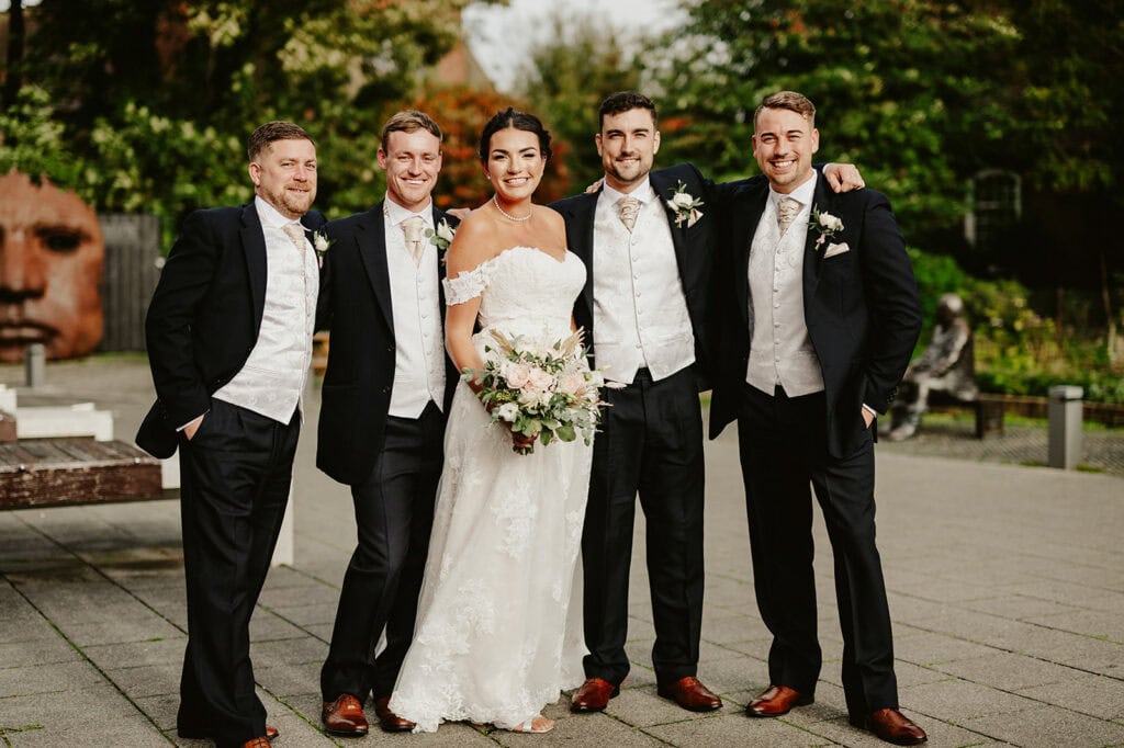 A bride in a lace wedding gown stands with four men in formal attire outdoors, smiling at the camera. The men are wearing dark suits with ivory vests. A large sculpture of a face is in the background.