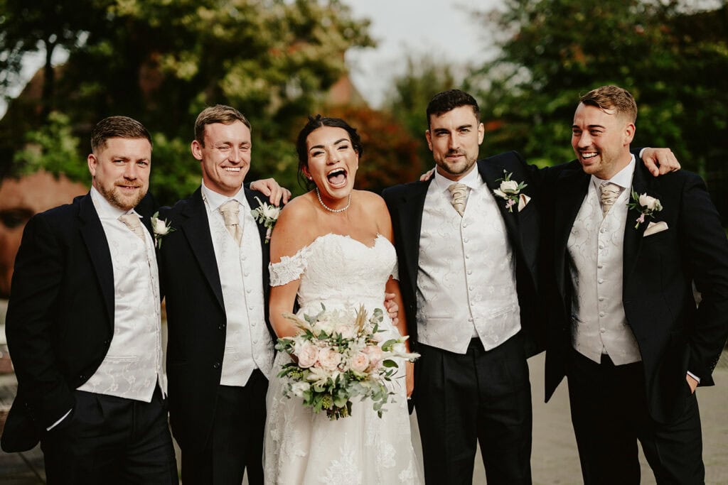 A bride in an off-shoulder wedding dress stands laughing in the center, holding a bouquet, surrounded by four men in matching suits and vests, all smiling outside with greenery in the background.