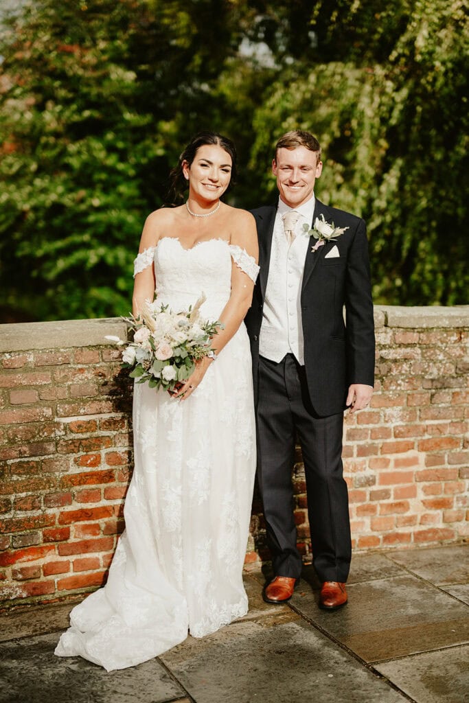 A smiling bride and groom stand outdoors, wearing a white lace wedding dress and a black suit respectively. They are positioned against a brick wall with greenery visible in the background.