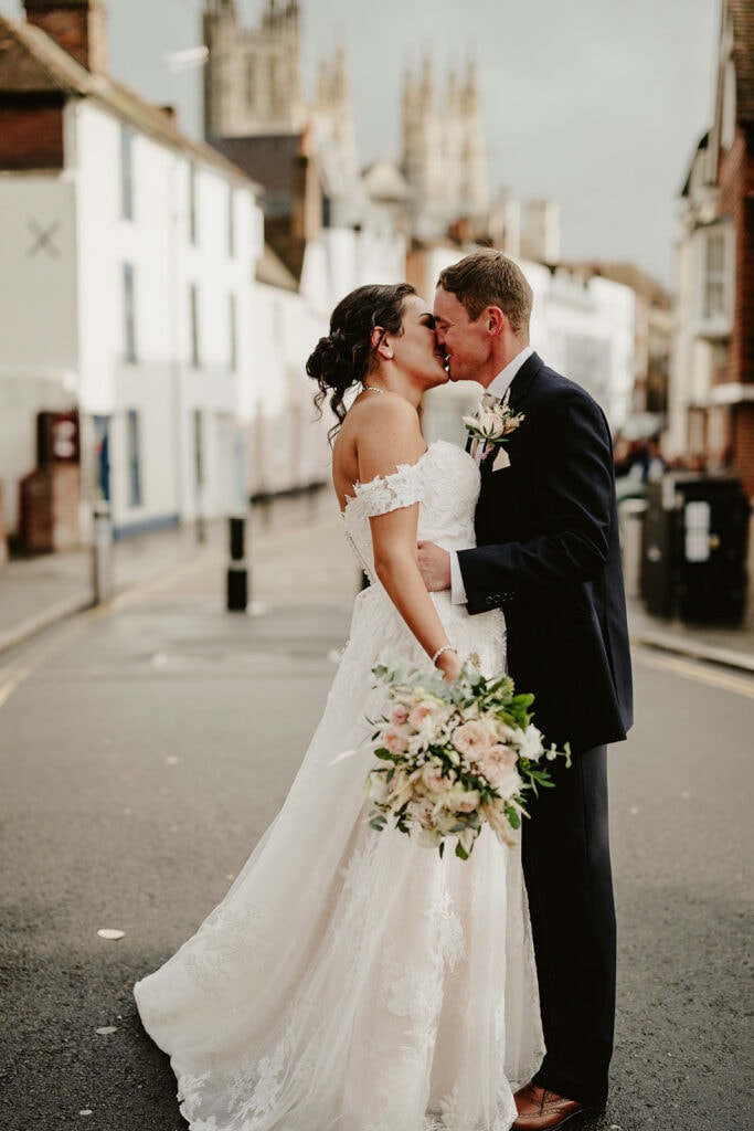 A bride and groom share a kiss on a street. The bride wears a white off-shoulder gown and holds a bouquet, while the groom is in a navy suit. A church is visible in the background.