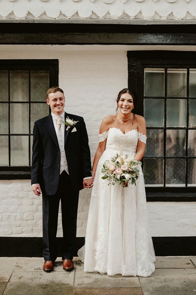 A bride and groom stand hand in hand in front of a white brick wall with black-trimmed windows. The bride holds a bouquet and wears a white off-shoulder wedding gown, while the groom is in a dark suit.